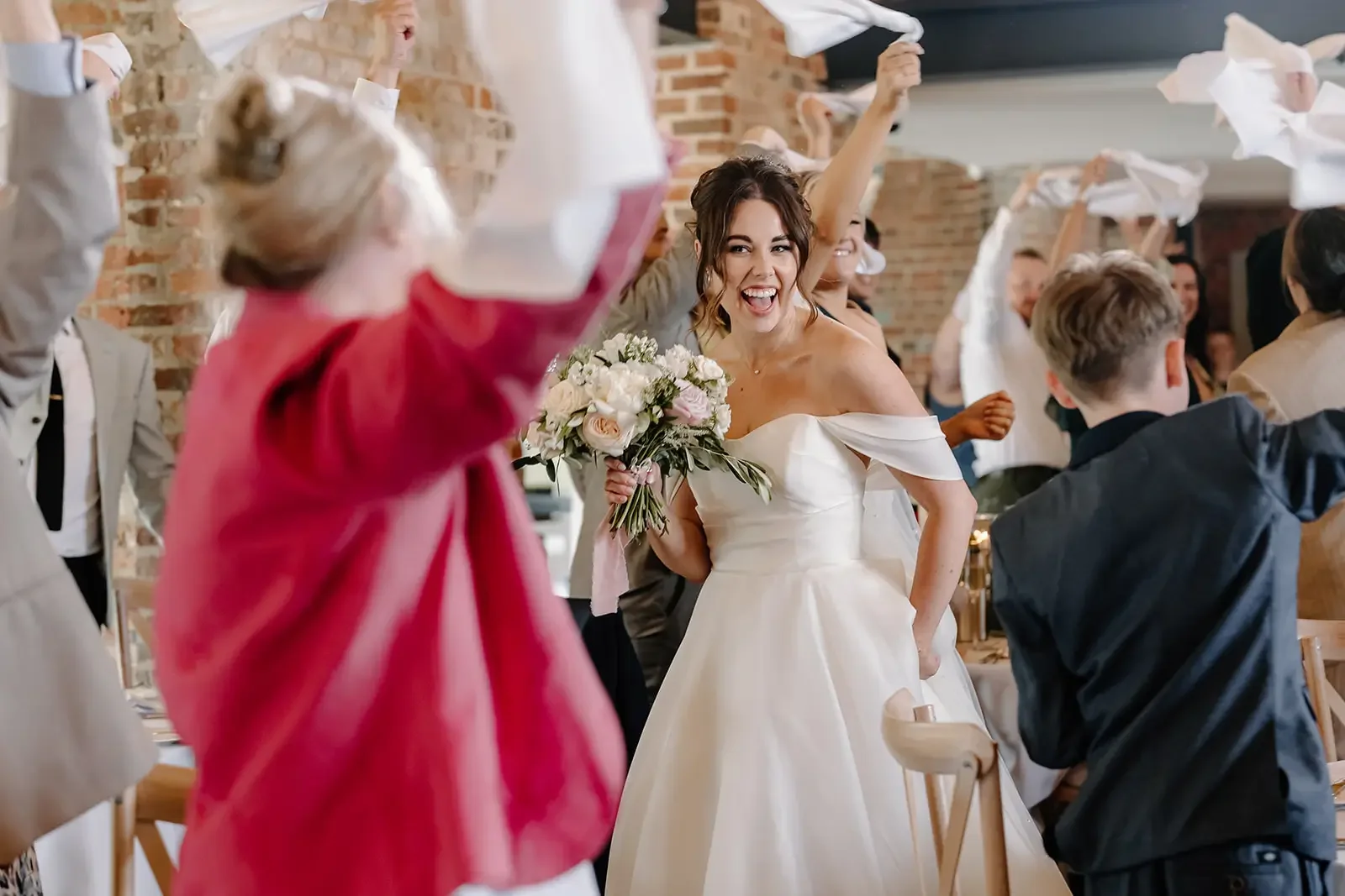 Bride celebrating with guests holding paper towels at a wedding reception, holding a bouquet of flowers and smiling.