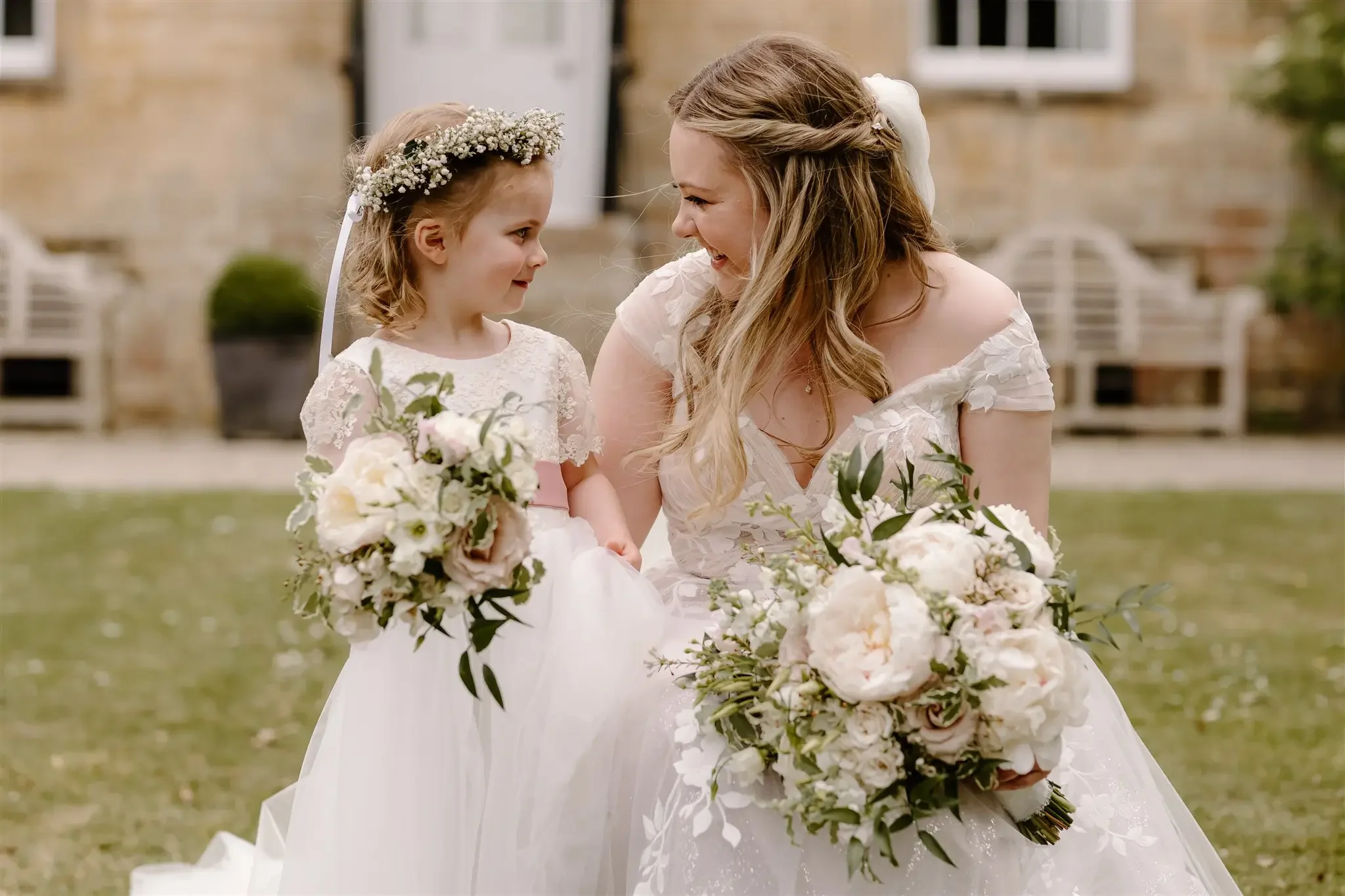 A bride and a young girl, possibly a flower girl, sharing an emotional moment outdoors at a wedding. The girl is holding a bouquet of flowers, wearing a flower crown, and the bride has a large bouquet and is smiling at the girl.