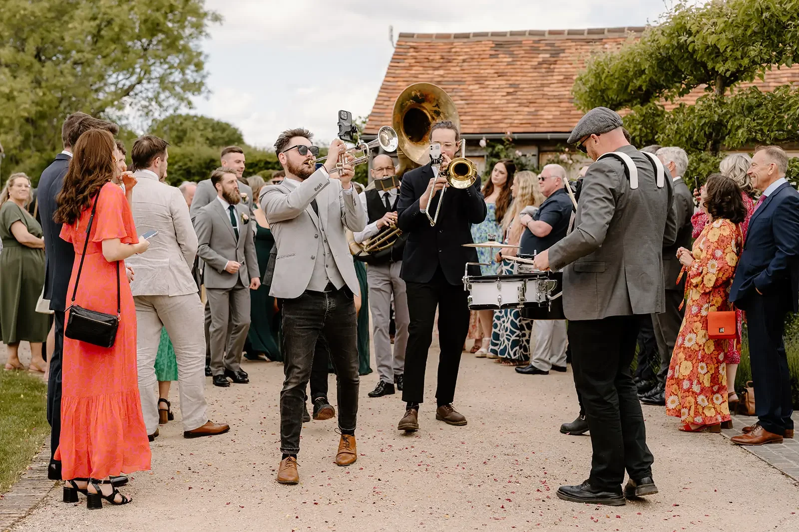 A group of musicians playing instruments in the middle of a crowd gathered outdoors, at a wedding or celebration. The musicians include a trumpet, a trombone, a saxophone, a tuba, and a drum.