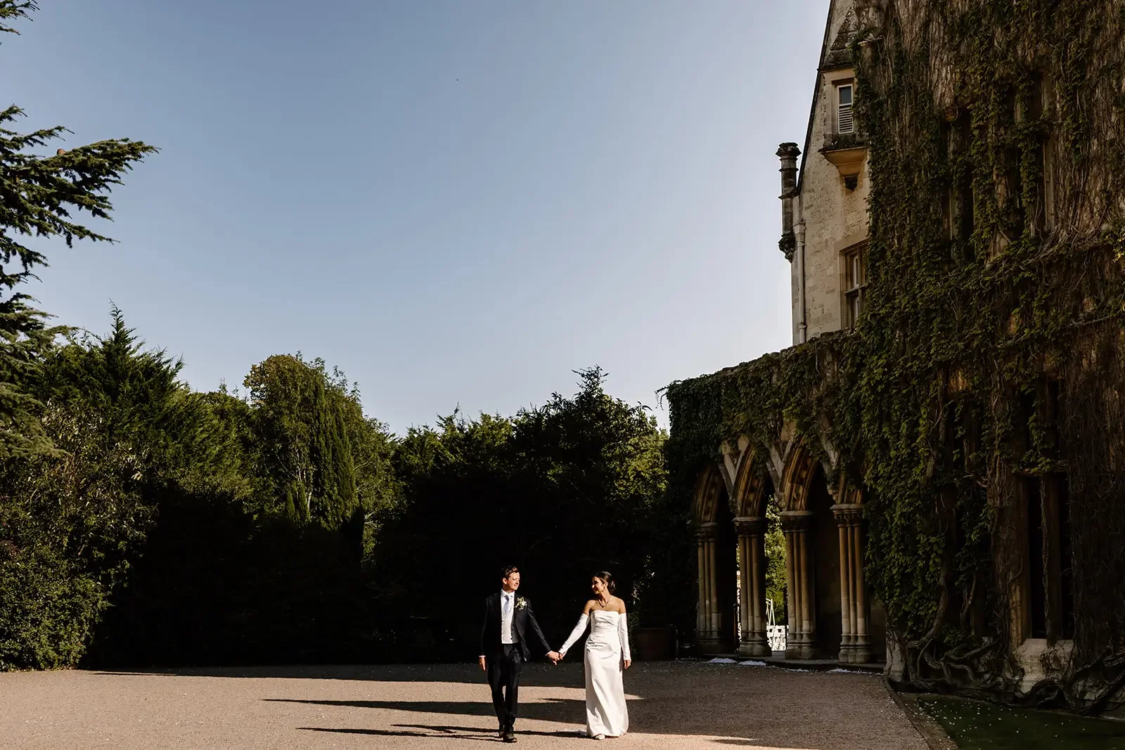 A bride and groom holding hands and walking outdoors near a historic building with arched windows and ivy on the walls.