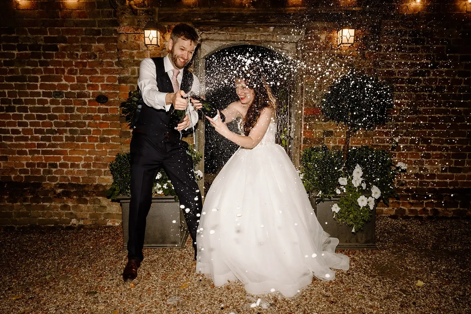 Bride and groom celebrating wedding with champagne, spray of champagne in the air, brick wall background, outdoor setting at night.
