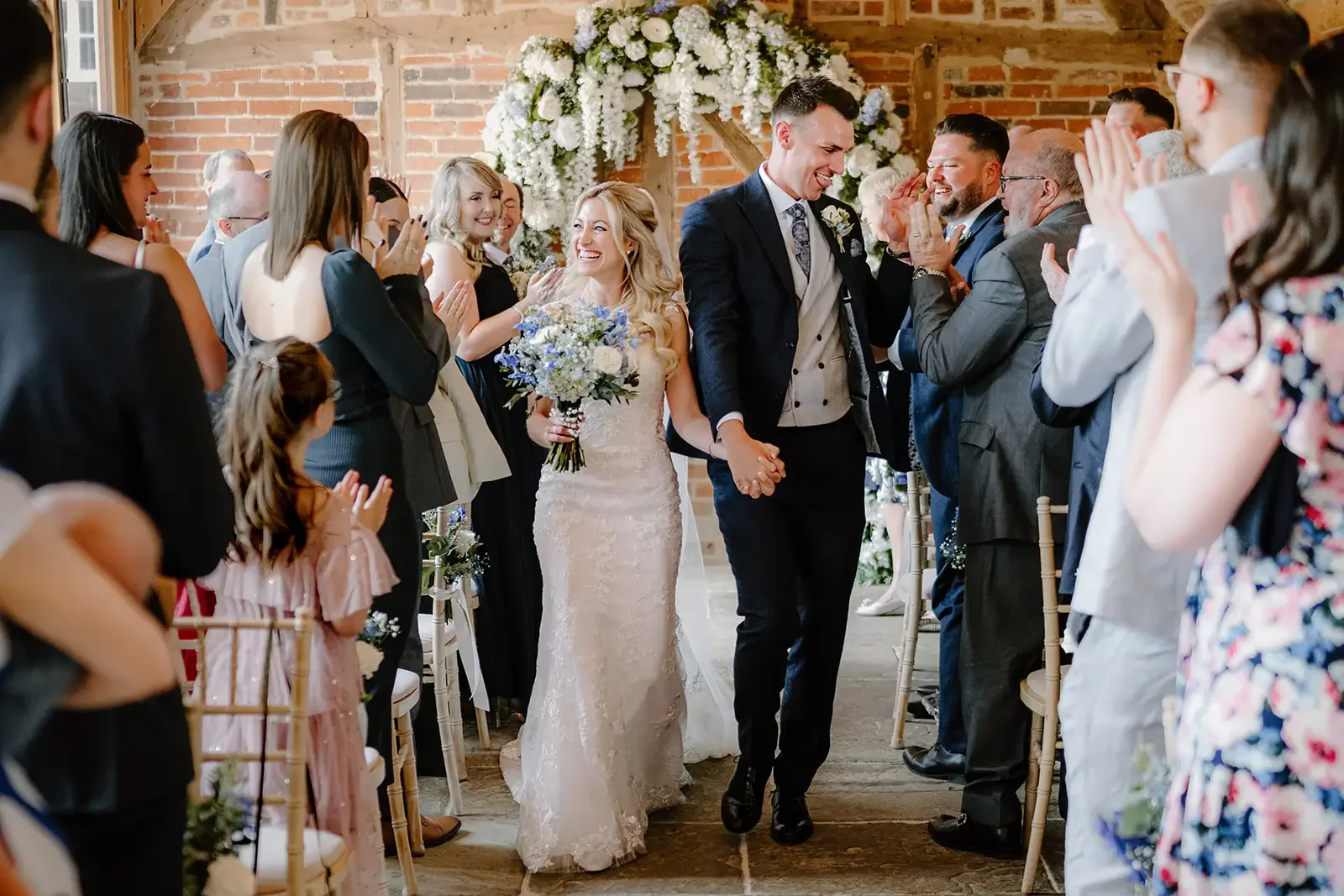 Bride and groom walking hand in hand through a celebratory aisle of friends and family during their wedding reception, with everyone clapping and smiling.