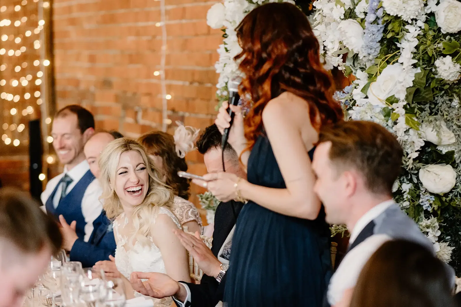 A woman with red hair is giving a speech at a wedding reception, holding a microphone and reading from notes. She is standing in front of a large floral arrangement with white flowers. The bride and groom are seated at a decorated table, smiling and 