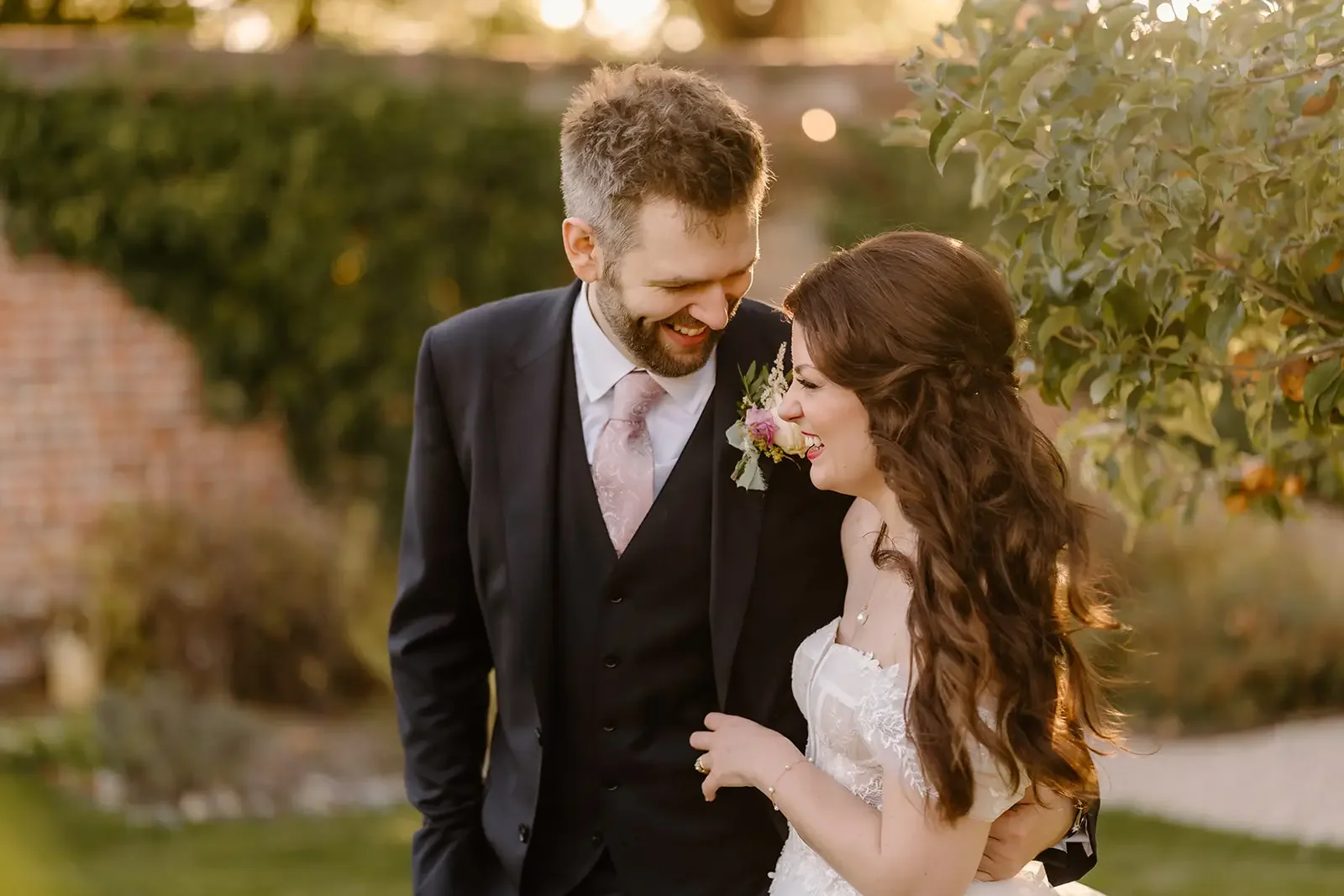 A newlywed couple smiling and leaning towards each other outdoors during sunset, with a blurred background of trees and a brick wall.