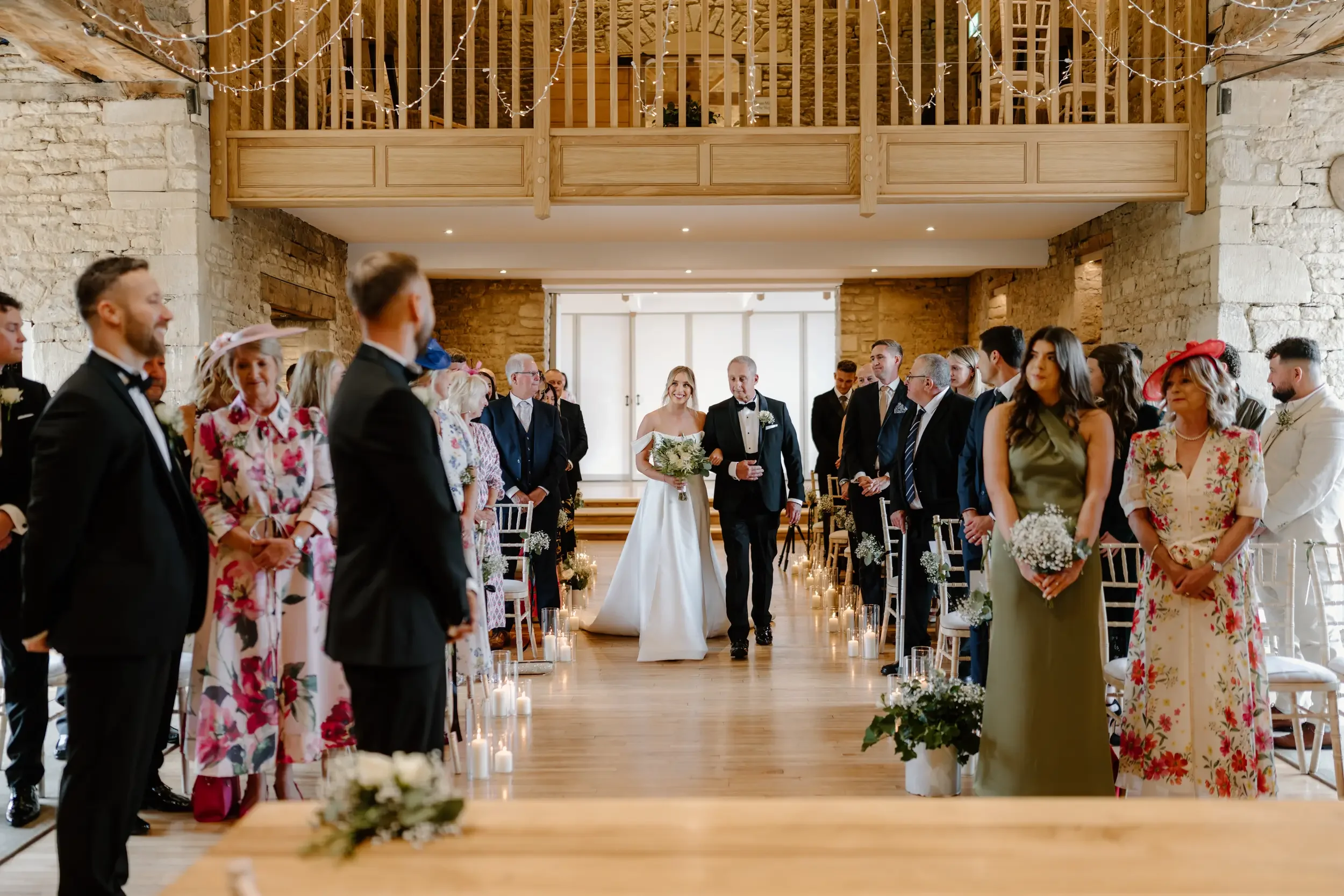 Wedding ceremony inside a rustic venue with stone walls, where a bride in a white gown is walking down the aisle escorted by an older man, possibly her father. Guests in formal attire are standing and watching the aisle, with some women wearing flora