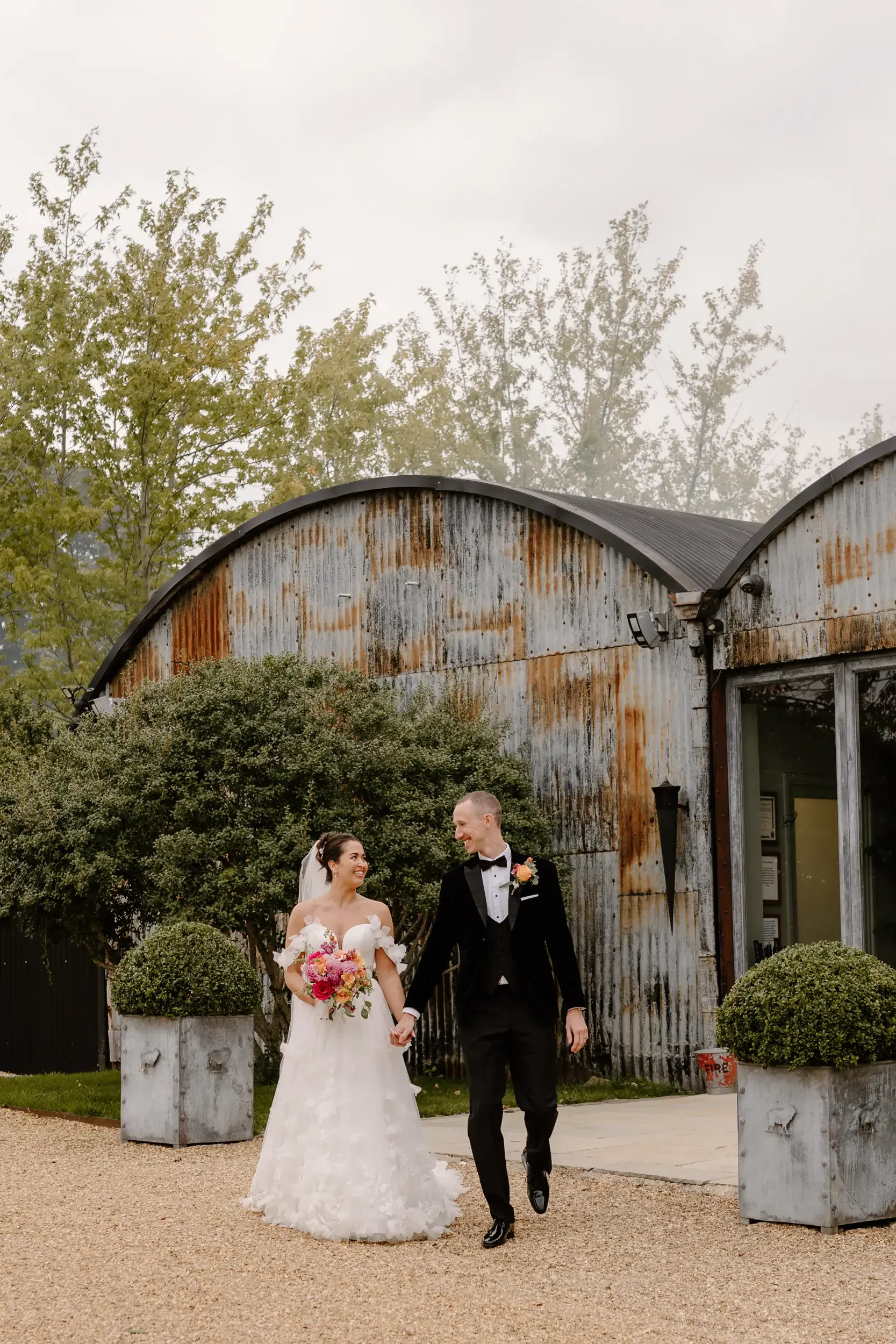 A newlywed couple in wedding attire walking hand-in-hand outdoors, with the bride in a white wedding gown holding a bouquet and the groom in a black tuxedo, in front of a rustic, weathered building surrounded by greenery.