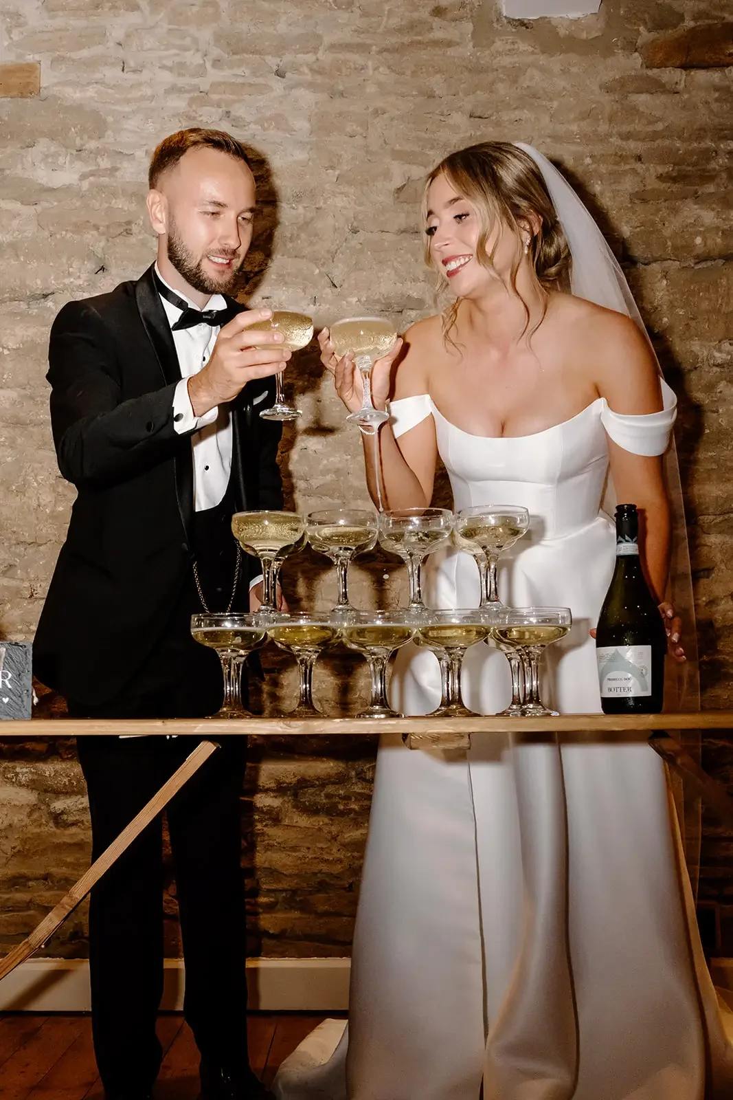 Bride and groom in wedding attire clinking champagne glasses to celebrate with a champagne tower against a brick wall.
