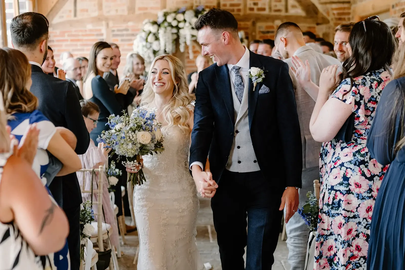 Bride and groom holding hands and smiling during their wedding ceremony, surrounded by guests clapping and celebrating indoors with a brick wall and floral decor in the background.