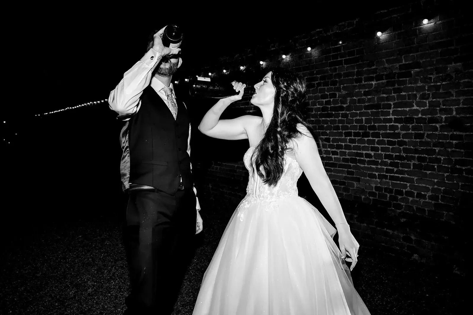 Bride and groom at a wedding, with the groom drinking from a bottle and the bride flexing her arm in a playful manner, against a brick wall background at night.