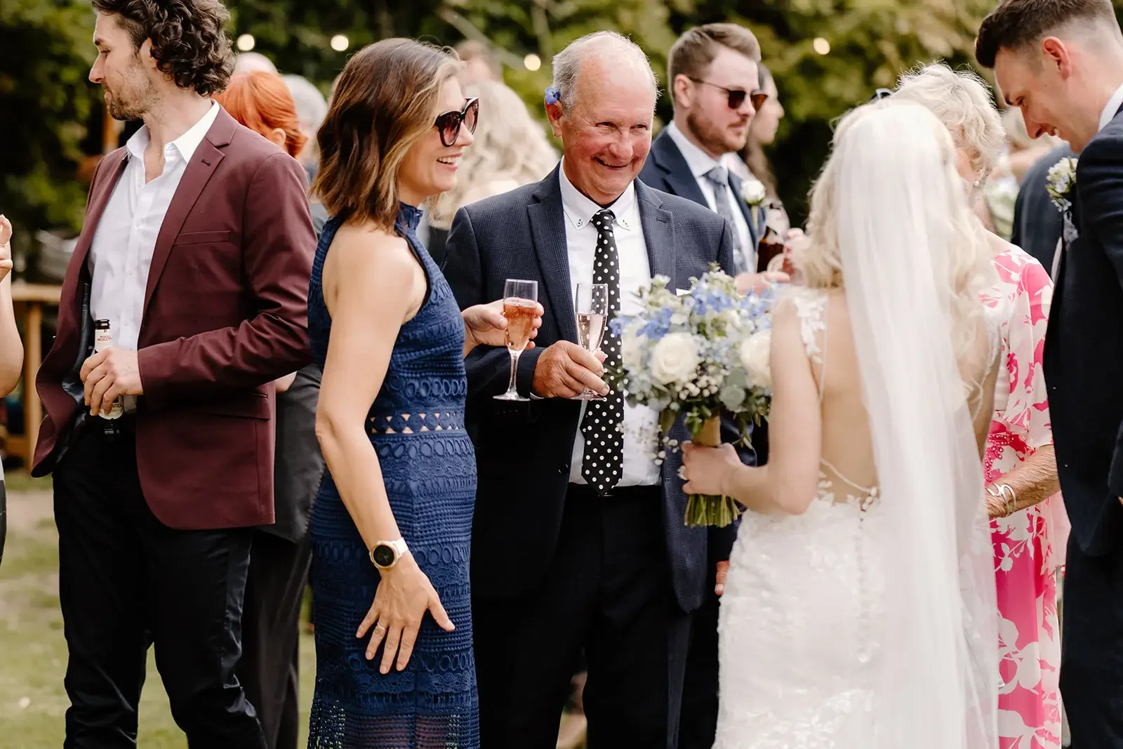 Group of wedding guests outdoors, talking and holding drinks, with a bride and groom in the background.