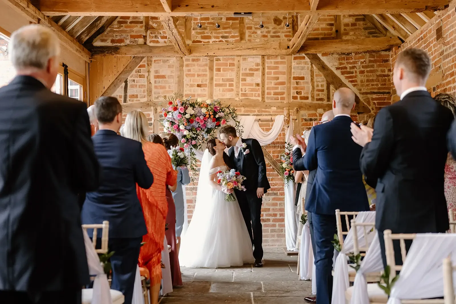 A bride and groom share a kiss at their wedding altar decorated with a large flower arrangement, as wedding guests stand and clap in a rustic brick venue.