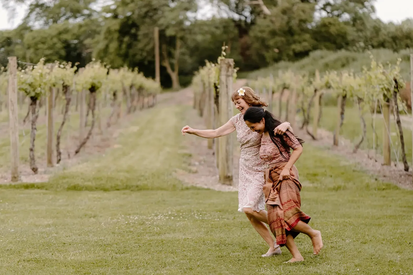 Two women joyfully running and laughing in a vineyard with grapevines in the background.