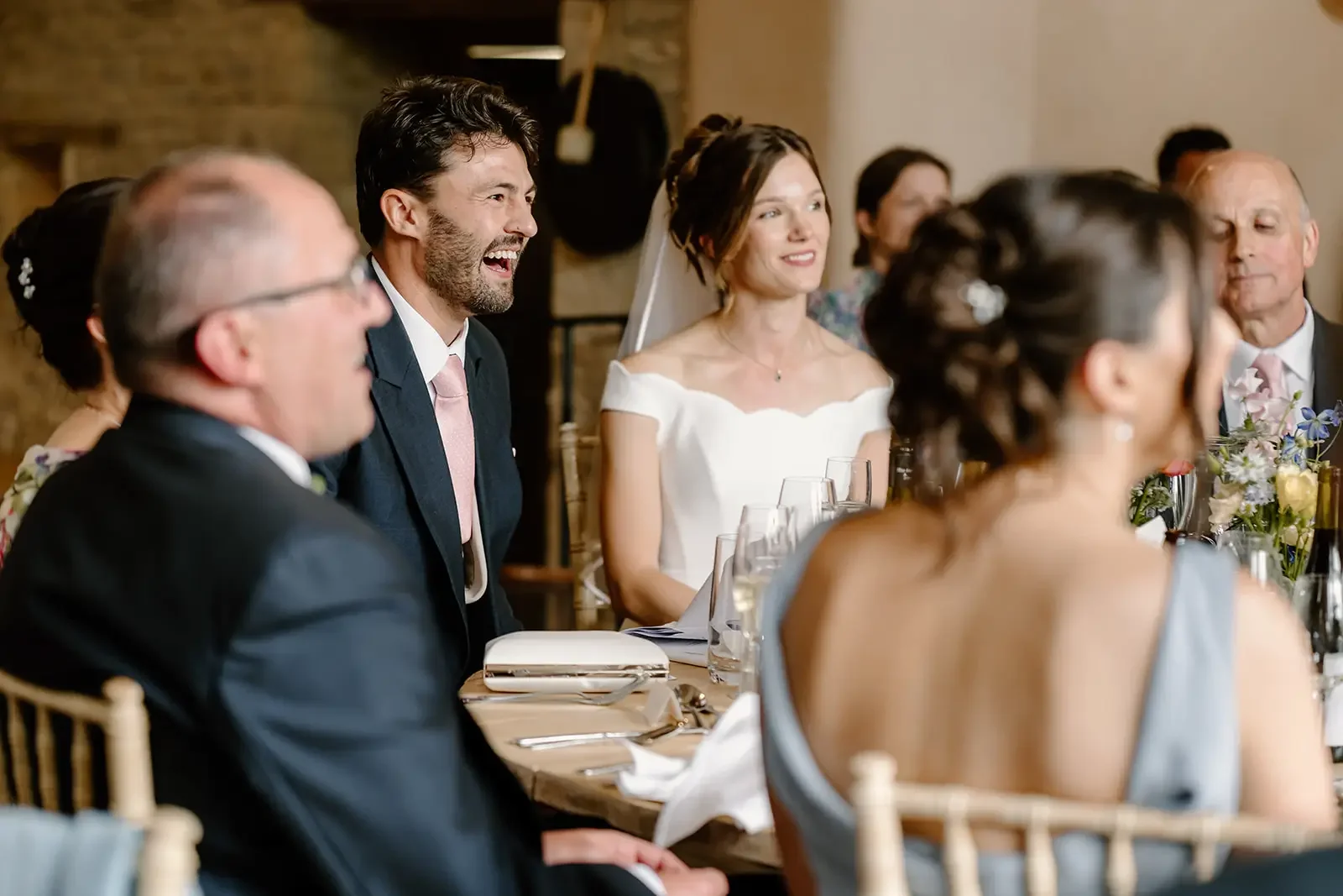 People at a wedding reception sitting at a table, smiling and laughing.