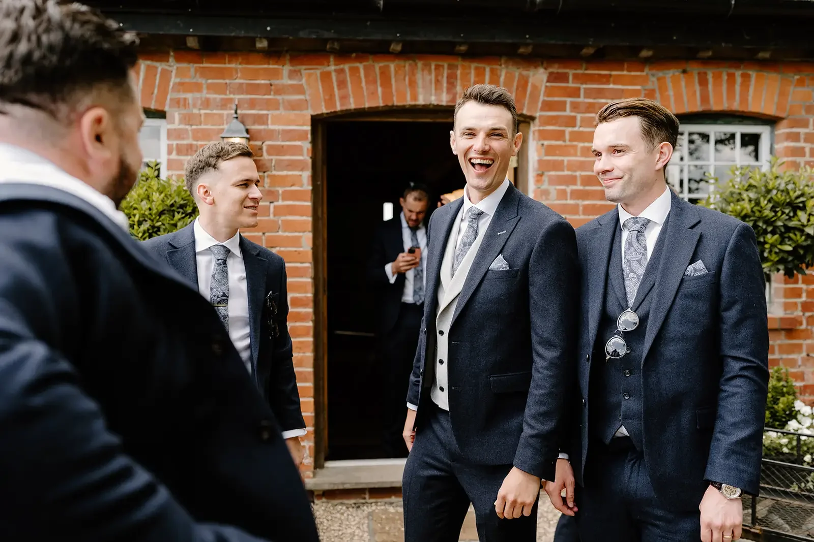 Men in suits standing outside a brick building, smiling and talking, one man in the background looking at his phone.