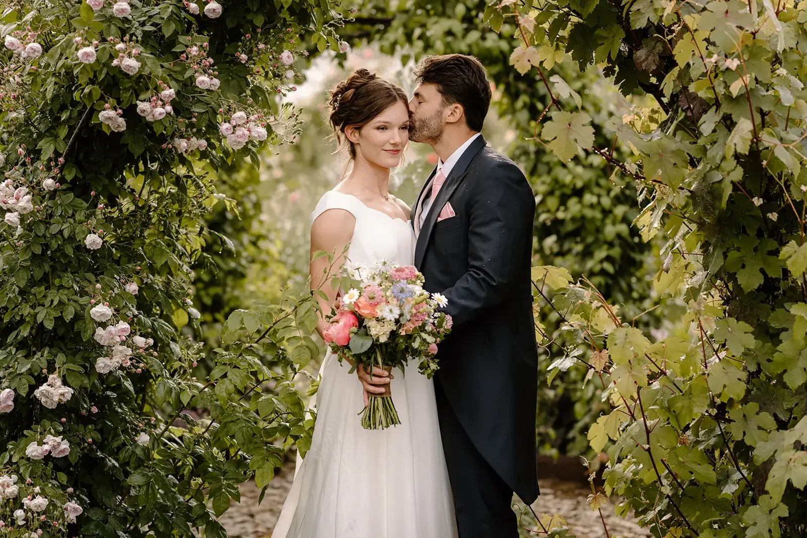 A bride and groom sharing a kiss outdoors among green foliage and blooming pink roses. The bride holds a colorful bouquet of flowers and wears a white dress, while the groom is in a dark suit with a pink tie and pocket square.