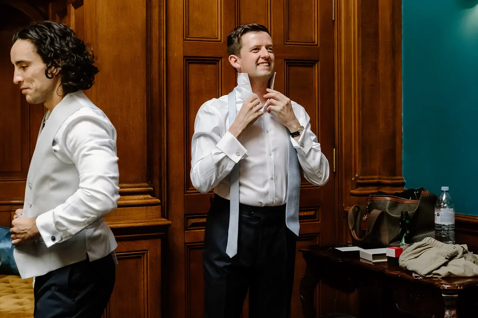 Man in white dress shirt with suspenders adjusting collar in room with wooden panels, table with water bottle, books, and bags.
