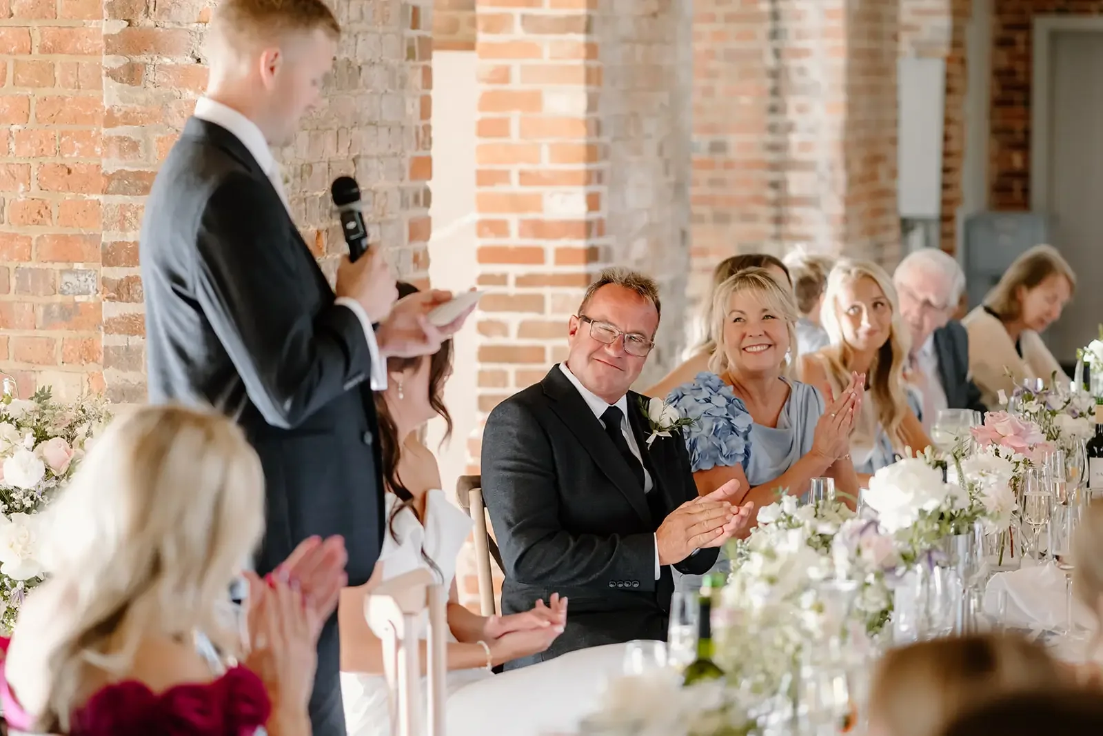 A man in a black suit gives a speech at a wedding reception which a group of guests, including a woman with blonde hair and a dark suit, are listening to and applauding.