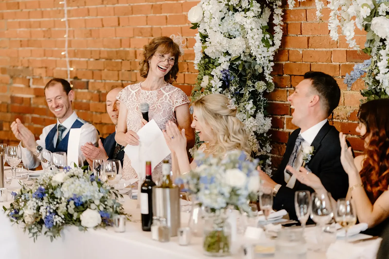 A woman giving a speech at a wedding reception, standing with a microphone and sheets of paper, surrounded by seated guests clapping and smiling, with floral decorations and a brick wall in the background.