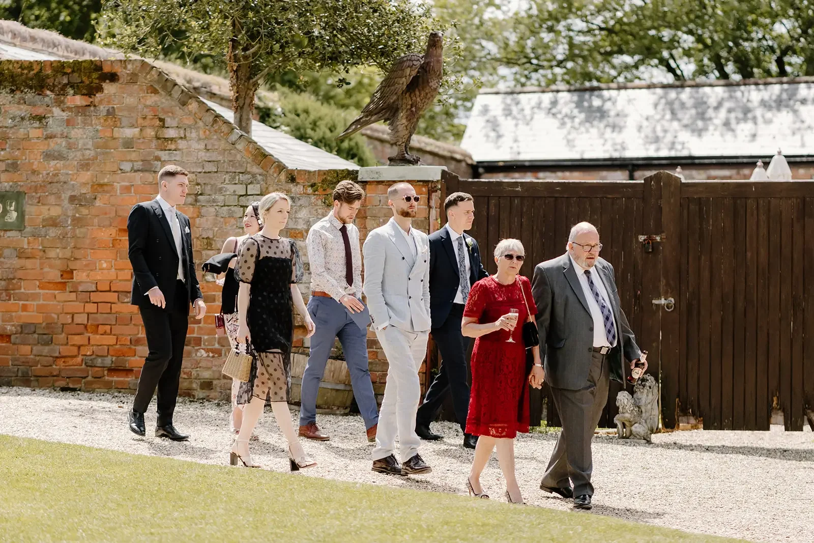 A group of people dressed in formal attire walking outdoors, with a brick wall, a wooden fence, and a large statue of an eagle in the background.