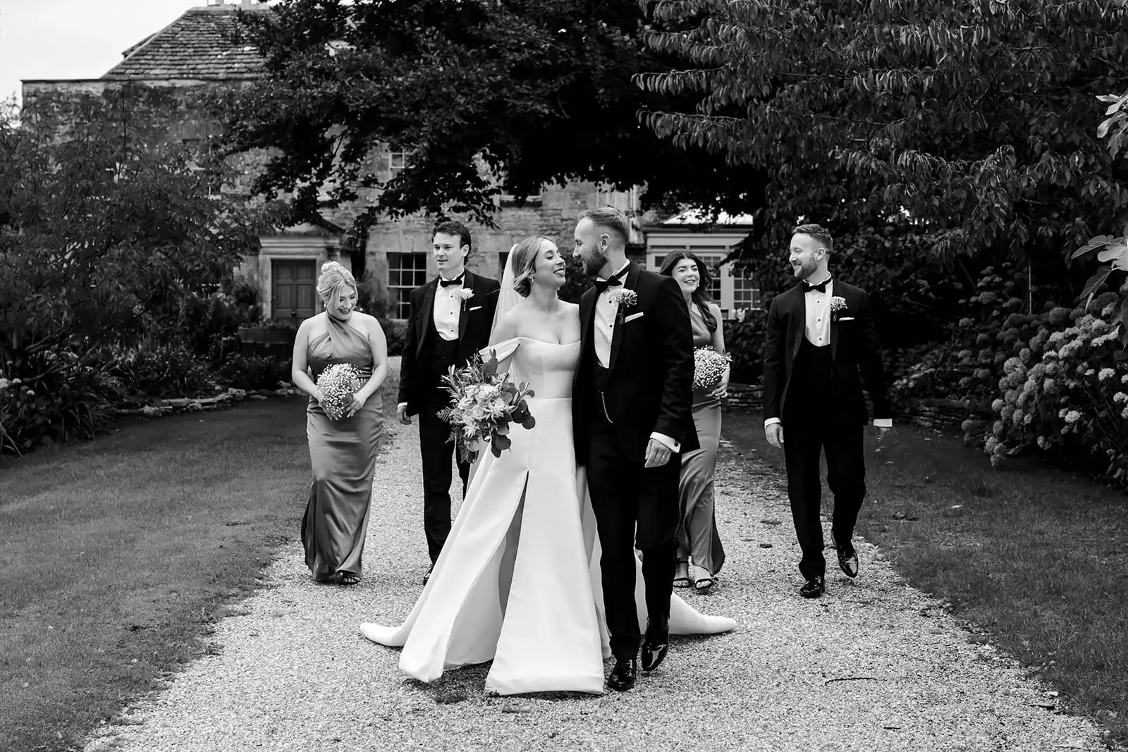 Black and white wedding photo of a bride and groom walking on a gravel path, surrounded by four wedding party members, with trees and a house in the background.