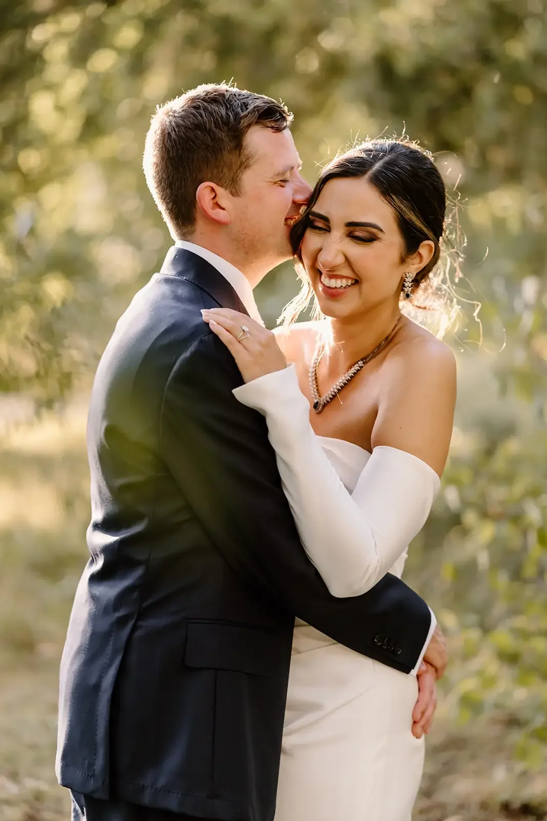 A newlywed couple sharing a joyful moment outdoors, with the groom playfully kissing the bride's cheek, in a sunlit natural setting.