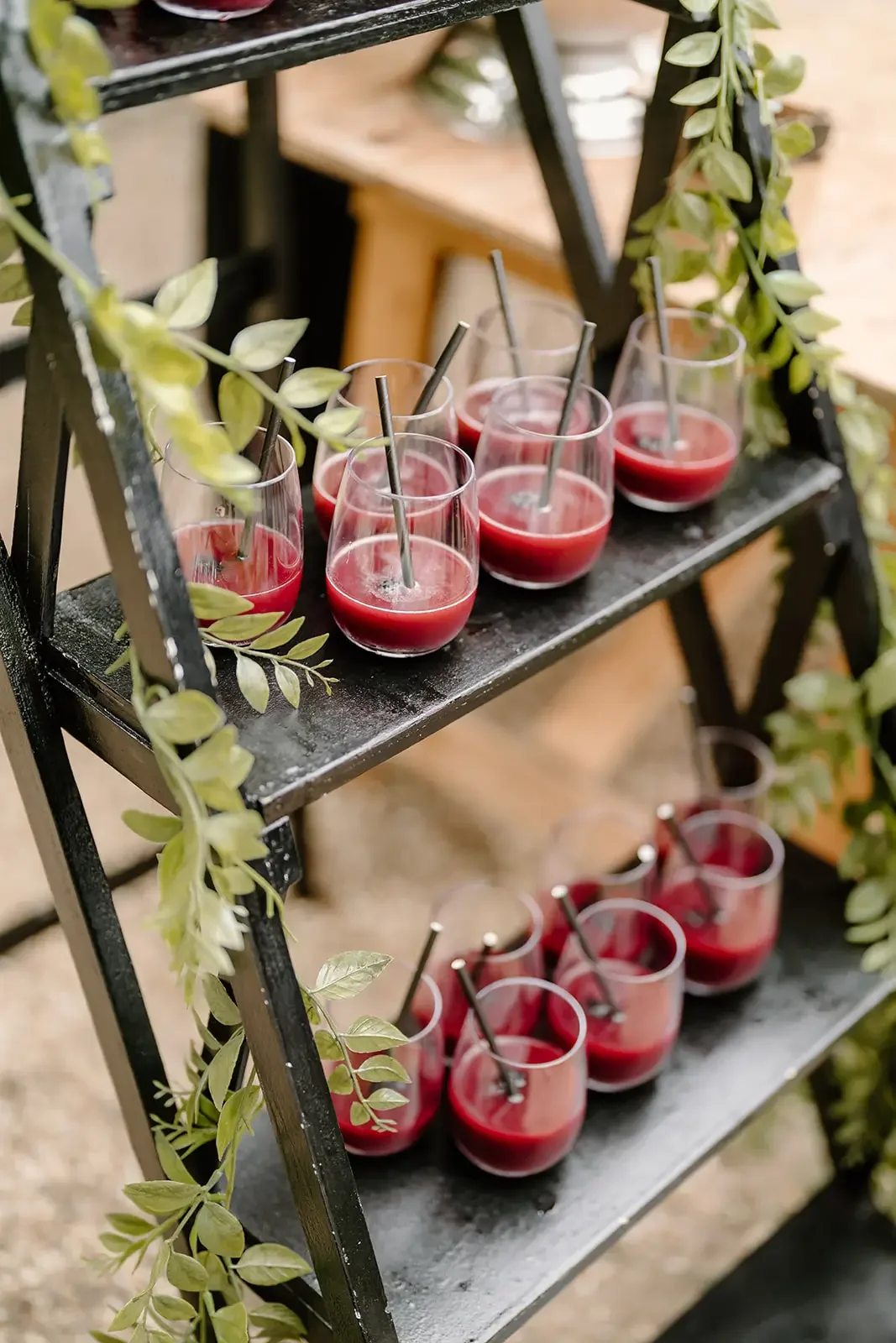 Two-tier black metal serving stand with green leaves draped over it, holding small glasses of red beverage with metal straws, set on a rustic surface.