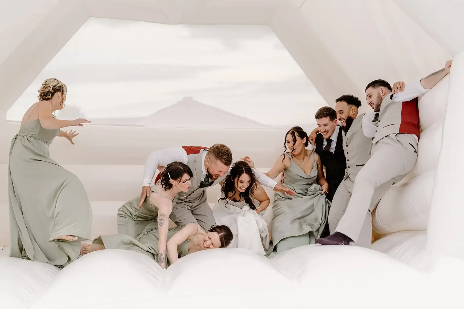 A group of people, including women in matching dresses and men in formal suits, engaging in a playful and joyous pillow fight on a large, fluffy white bed in a bright room with a large window showing an outdoor view.
