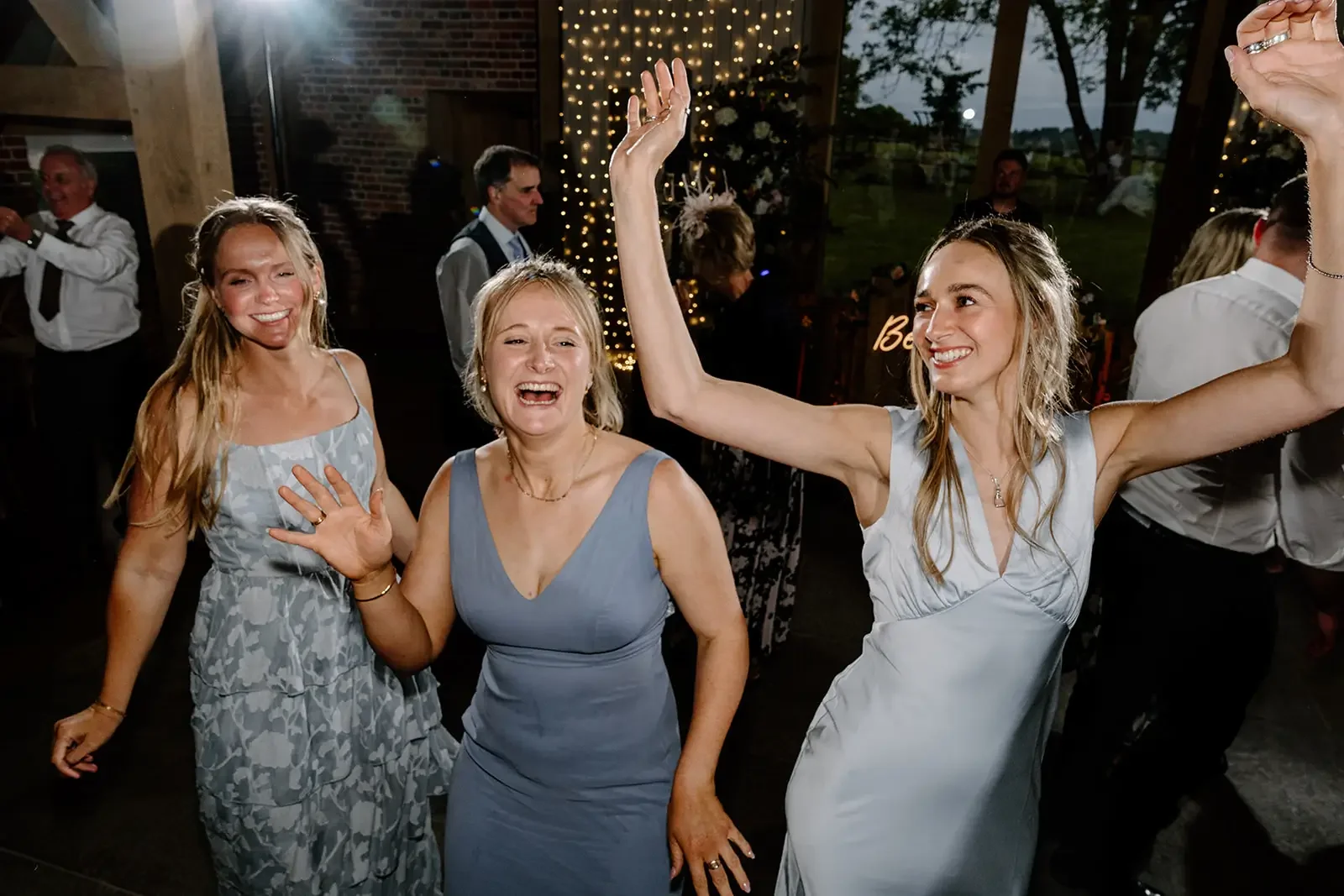 Three women in cocktail dresses dancing and smiling at a celebration or party, with people in the background, decorated with string lights and a dark artificial indoor setting.