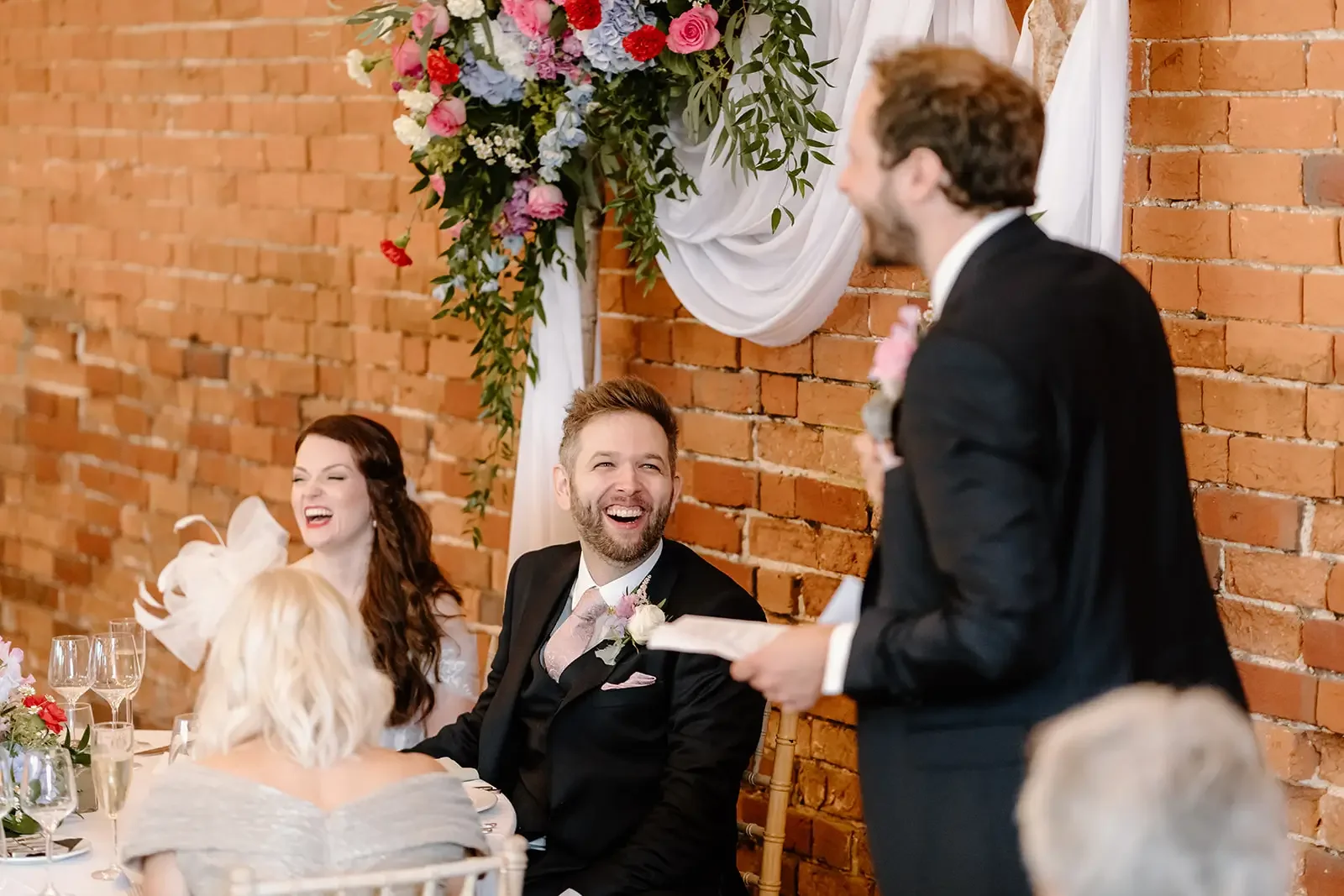 A wedding reception with guests seated at a table decorated with flowers, listening to a man giving a speech while a woman laughs and a man smiles in the background. The background features a brick wall with a floral and fabric decoration.