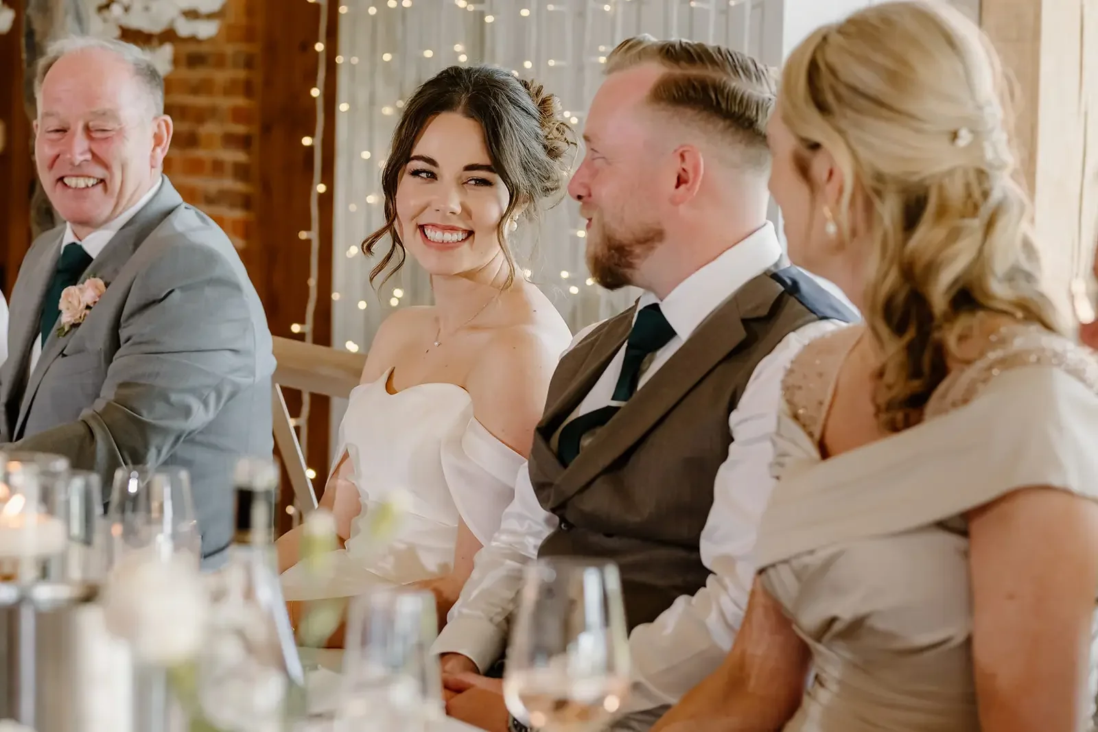 A group of people seated at a wedding reception table, including a smiling bride in a white strapless dress, a groom in a brown vest, an older man in a gray suit with a pink boutonniere, and a woman in a light-colored dress with lace details, next to