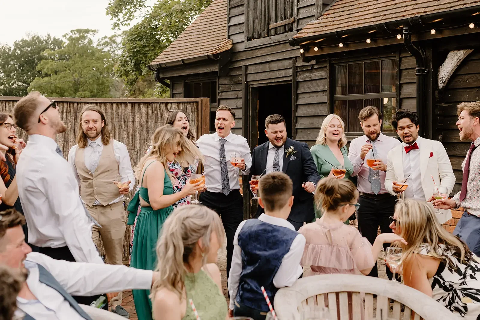 A group of people celebrating at an outdoor party, some are dancing and holding drinks, standing in front of a wooden building with string lights.