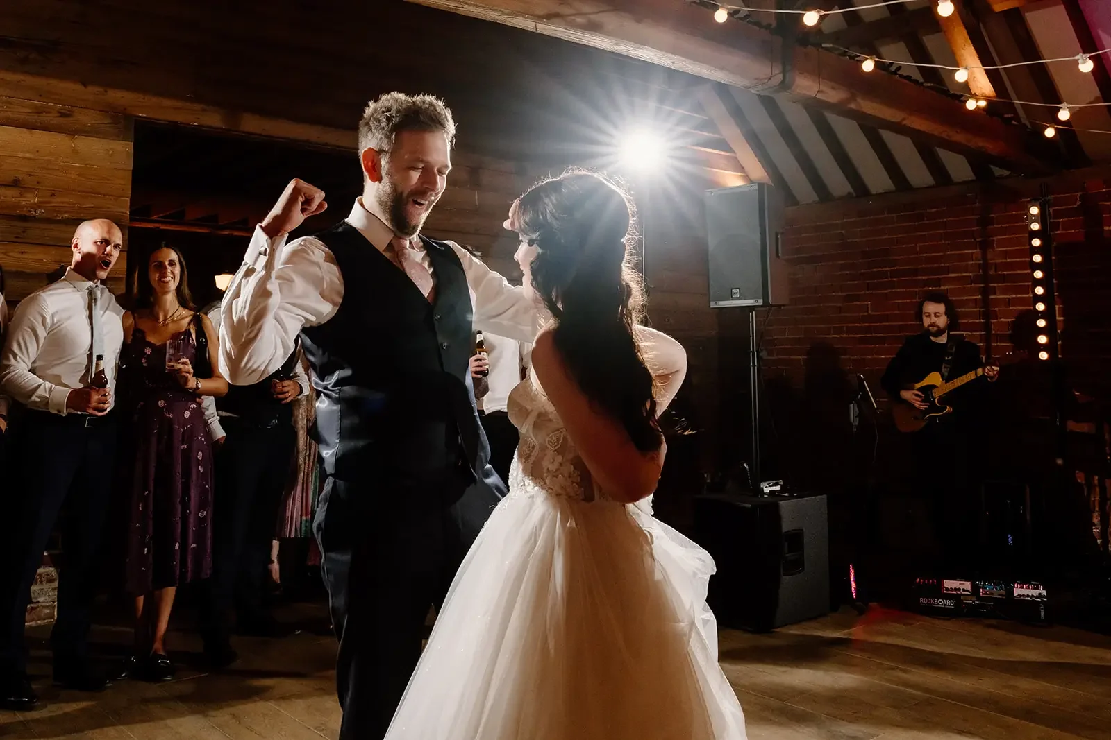A bride and groom dancing at their wedding reception, surrounded by guests, in a rustic venue with wooden walls and string lights, with a musician playing guitar in the background.