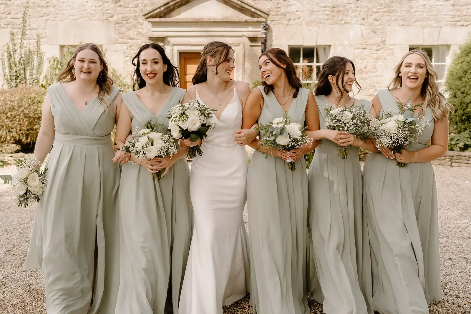 Bride and bridesmaids walking outdoors in front of a stone building, holding bouquets of white flowers, dressed in coordinated light gray dresses, smiling and chatting with each other.