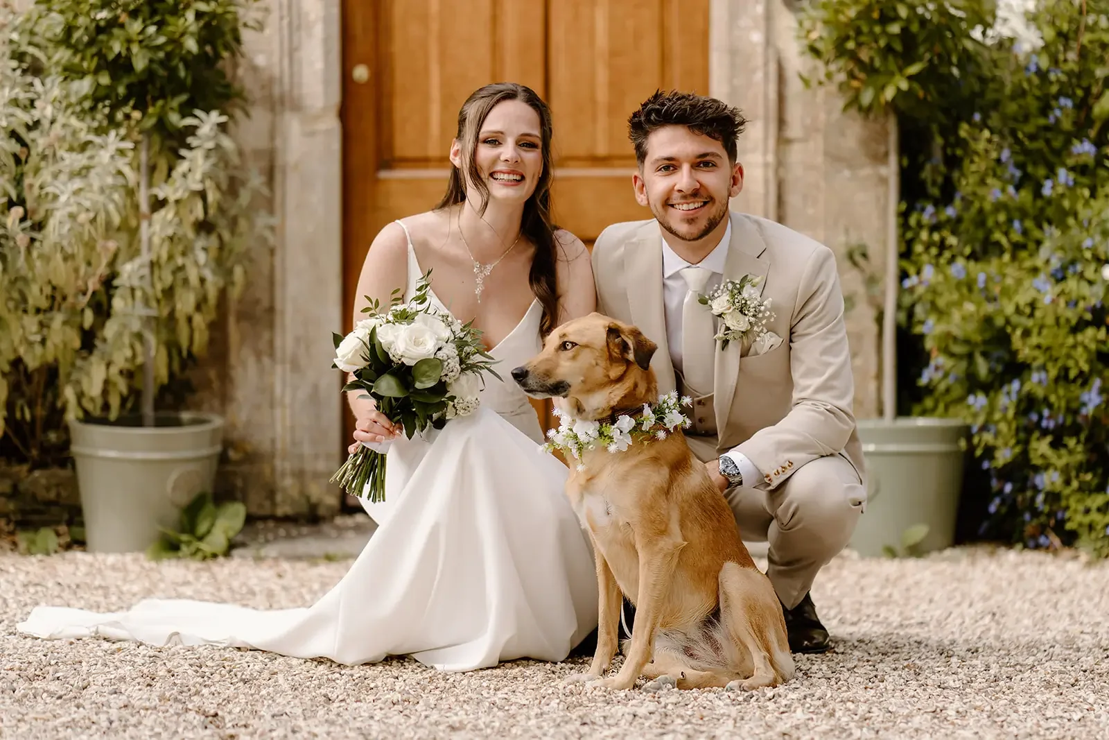 A bride and groom in wedding attire, kneeling beside a gold-colored dog wearing a floral collar, outdoors in front of a wooden door with greenery on either side.