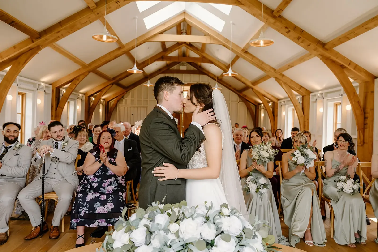 A bride and groom kiss during a wedding ceremony inside a wooden chapel, surrounded by seated guests who are clapping, smiling, and taking photos.