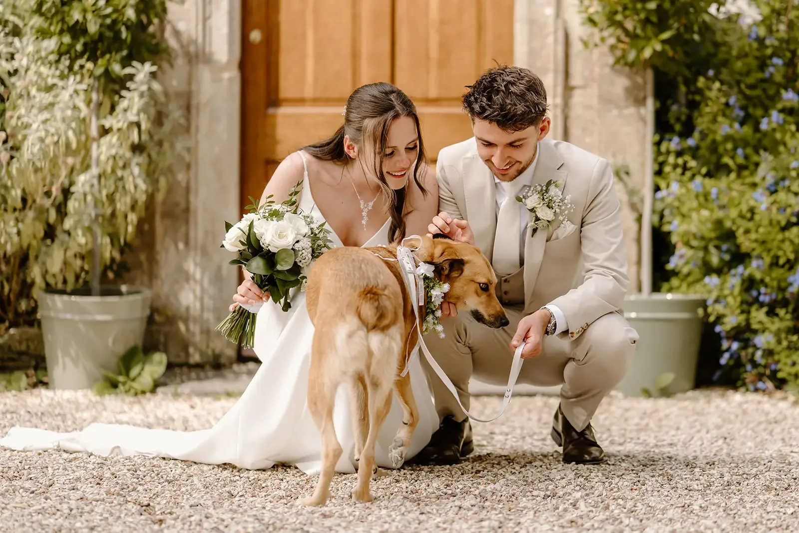 A bride and groom in wedding attire kneeling outdoors with a brown dog on a leash, both holding white flower bouquets, in front of a wooden door and greenery.