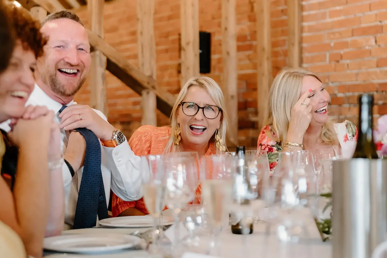 A group of adults sitting at a dinner table, laughing and enjoying a social gathering in a rustic setting with brick walls and wooden beams.