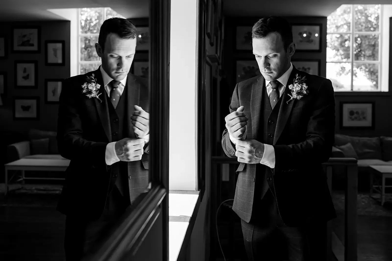 A man in a suit putting on a cufflink while looking into a mirror on both sides of the image. The scene is in a well-lit room with framed pictures on the wall and a window in the background.