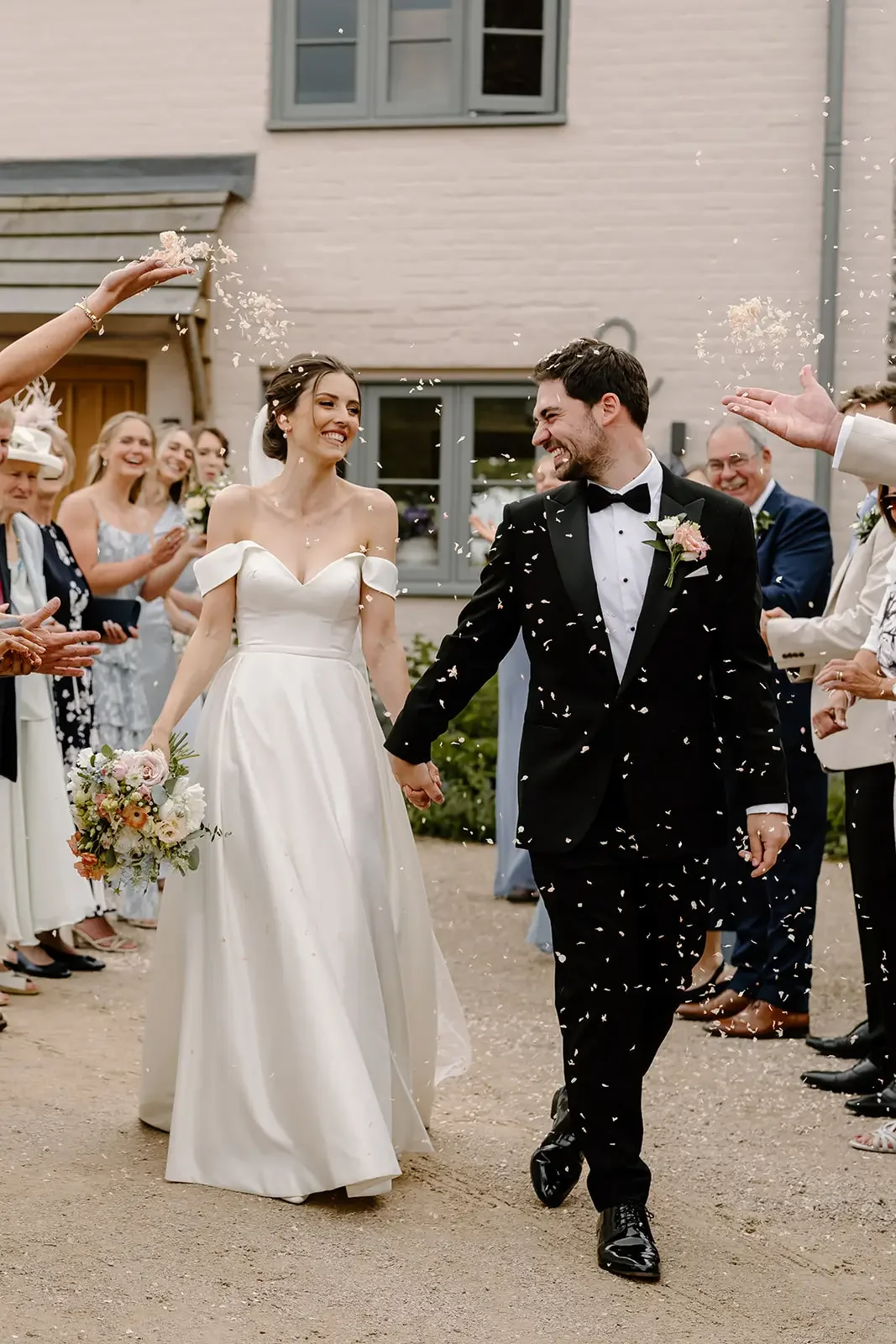 A bride and groom hold hands and walk through a celebration crowd, with confetti falling around them, smiling.
