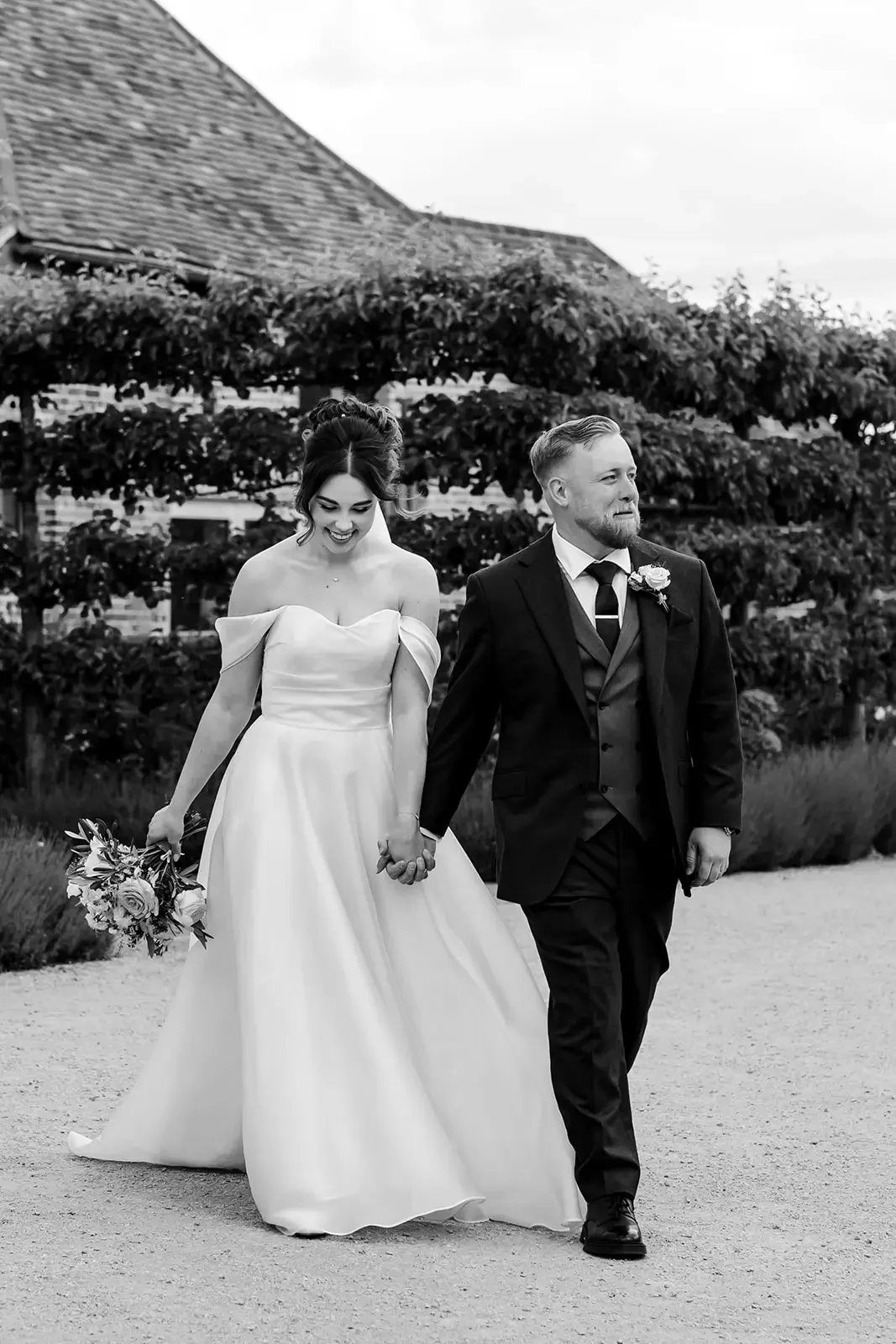 A bride and groom holding hands and walking outdoors on their wedding day, with the bride in a white gown holding a bouquet and the groom in a suit with a boutonniere, smiling and enjoying the moment.