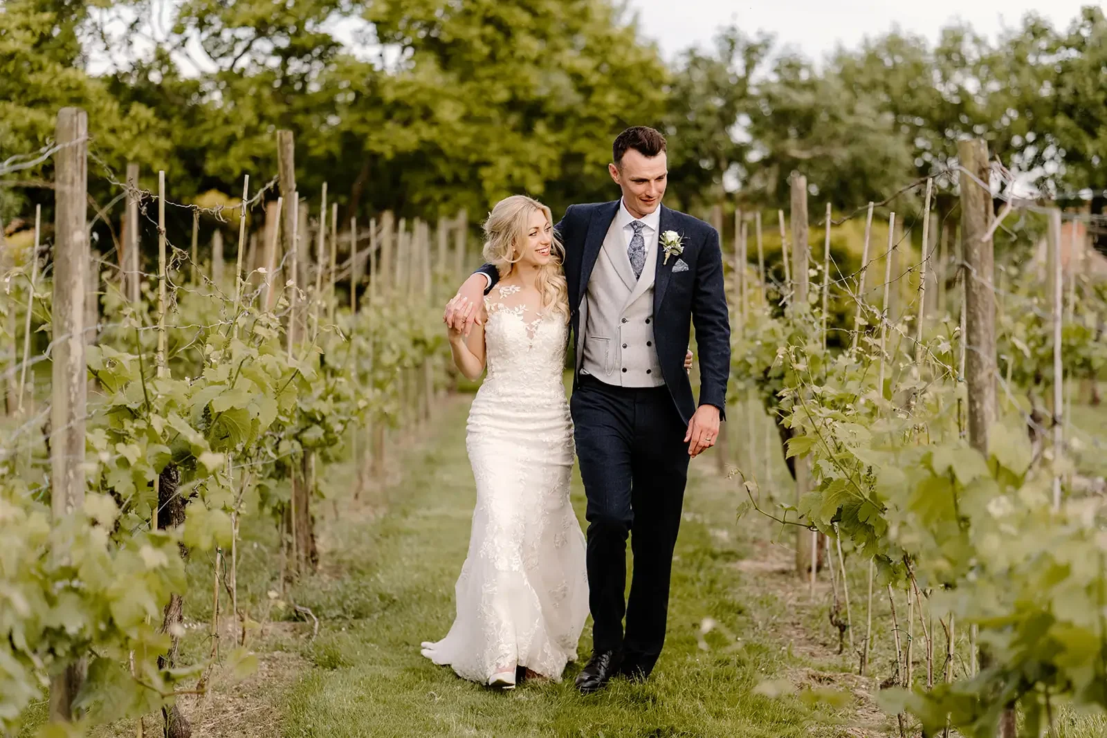 A bride and groom walking through a vineyard, smiling and holding each other.