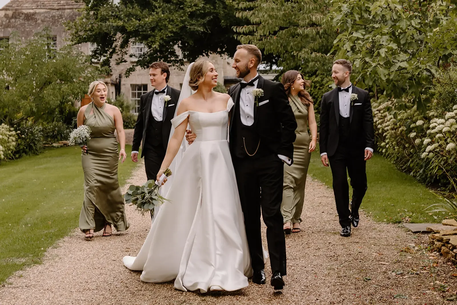 A wedding party walking outdoors on a gravel path with greenery and a stone building in the background. The bride and groom are smiling at each other, the bride in a white strapless wedding gown and the groom in a black tuxedo. Four other people, two