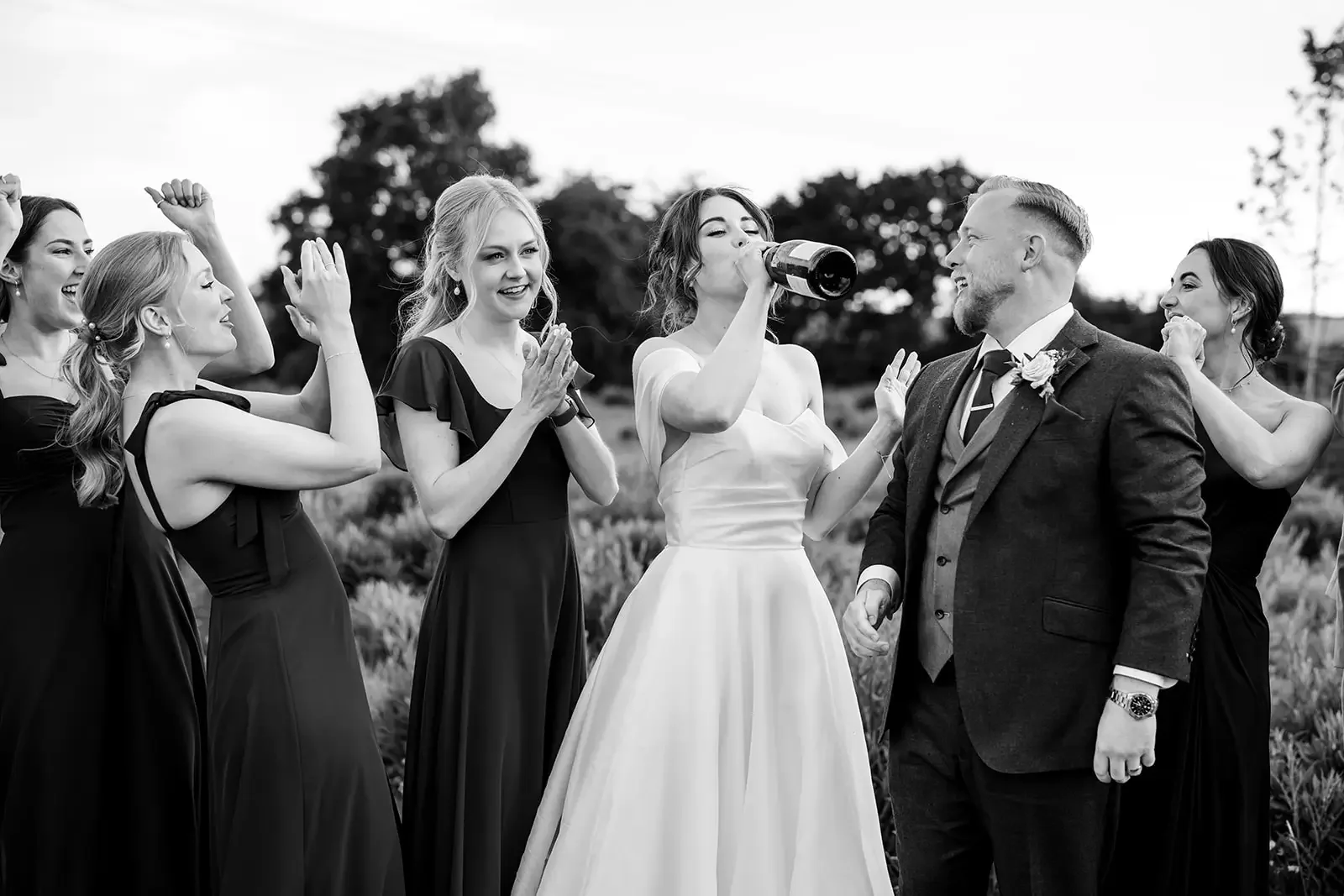 A bride in a white wedding gown drinks from a bottle while standing outside with a group of women and a man in a suit, celebrating at a wedding.