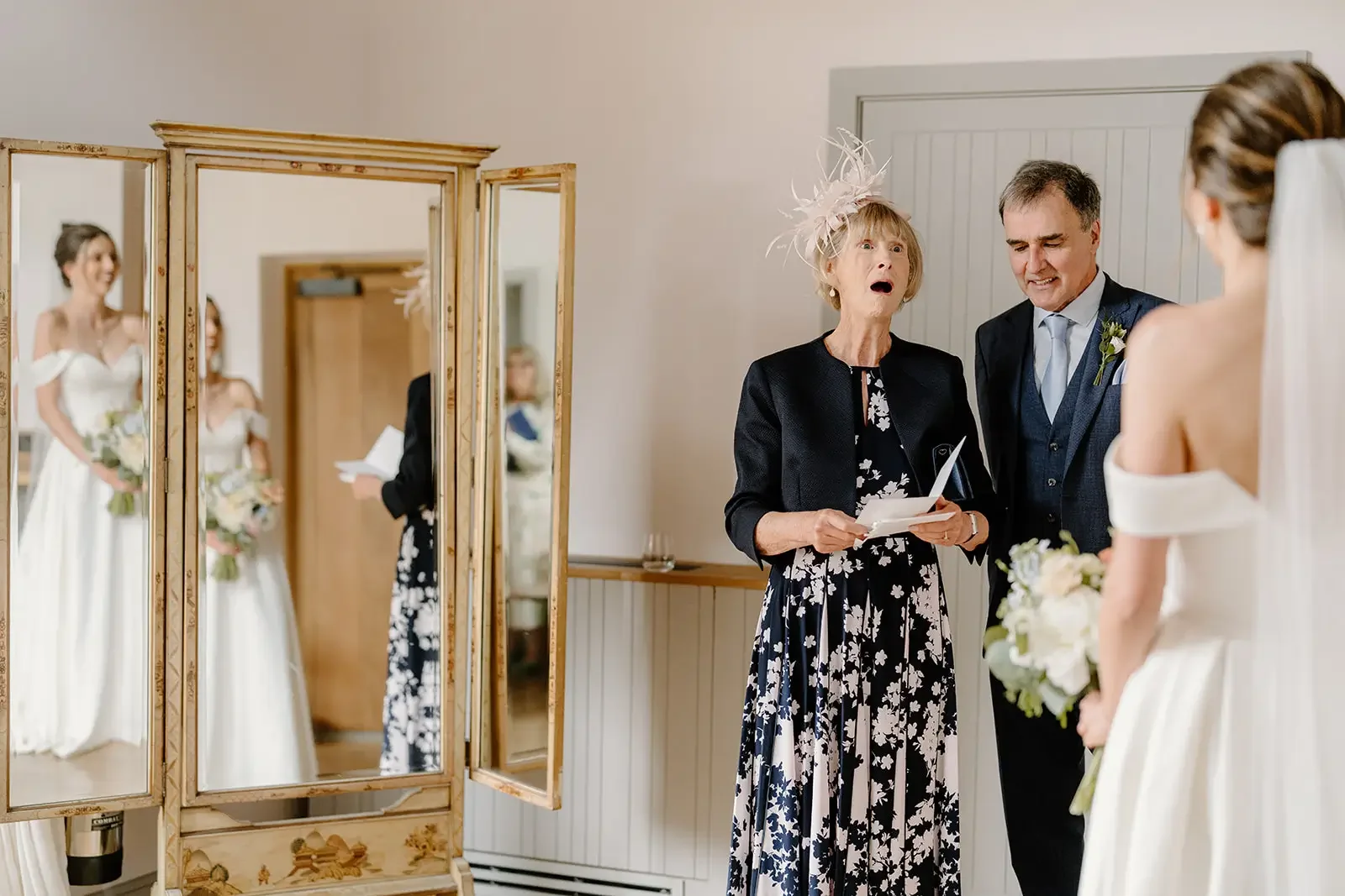 A bride in a white off-the-shoulder wedding dress holding a bouquet stands in front of a mirror. An older woman with a feathered hat and a floral dress appears to be reading vows or a speech, and a man in a suit is smiling beside her. The reflection 
