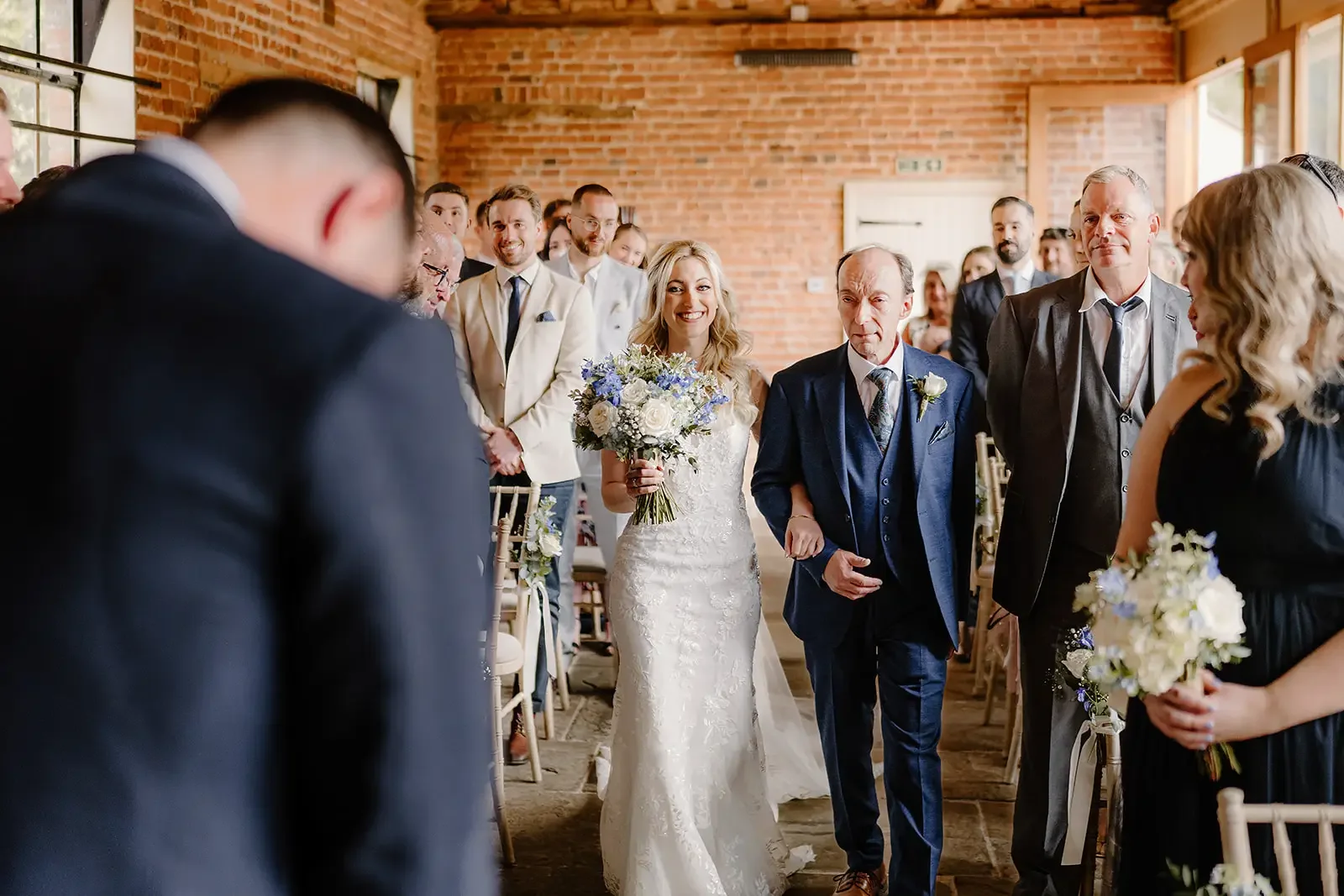 Bride walking down the aisle with her father during a wedding ceremony in a brick-walled venue, with guests watching and smiling.