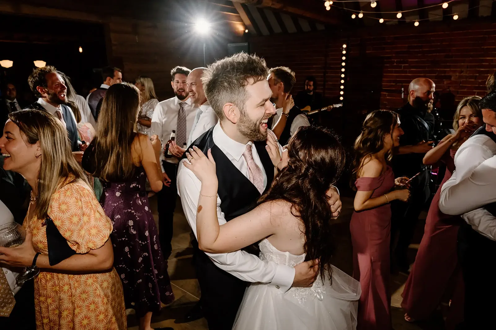 A group of people dancing and celebrating at a wedding reception inside a rustic venue, with a bride and groom in the center.