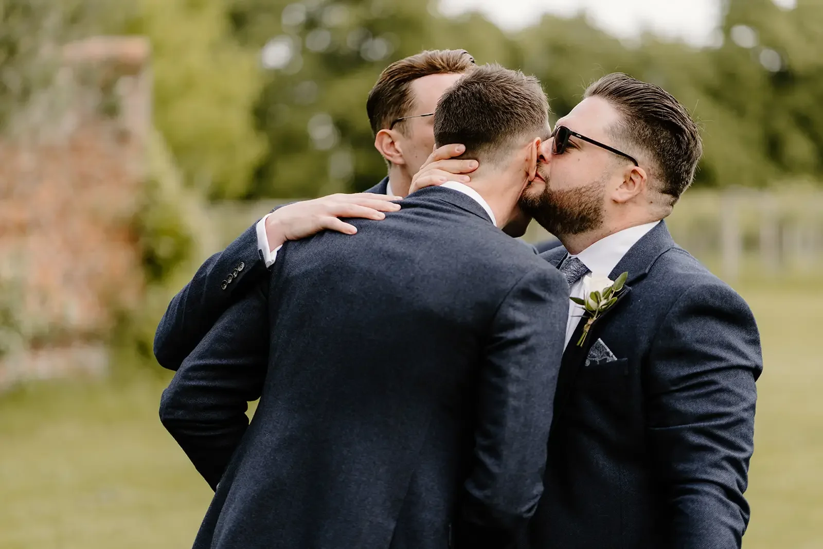 Three men in suits kissing and hugging outdoors during daytime, with blurred green trees in the background.