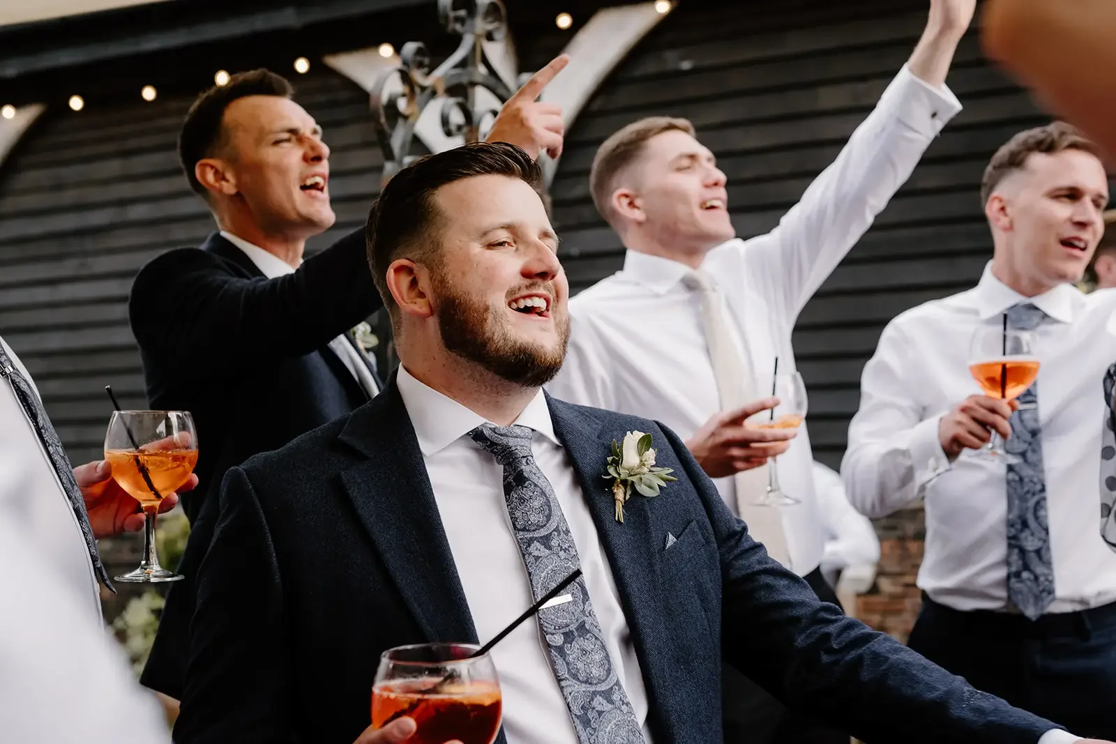 Group of men in formal attire celebrating at a wedding reception, some holding glasses of pink cocktails, with one man in a suit with a boutonniere smiling and others singing or talking with drinks in hand, against a dark wooden wall.