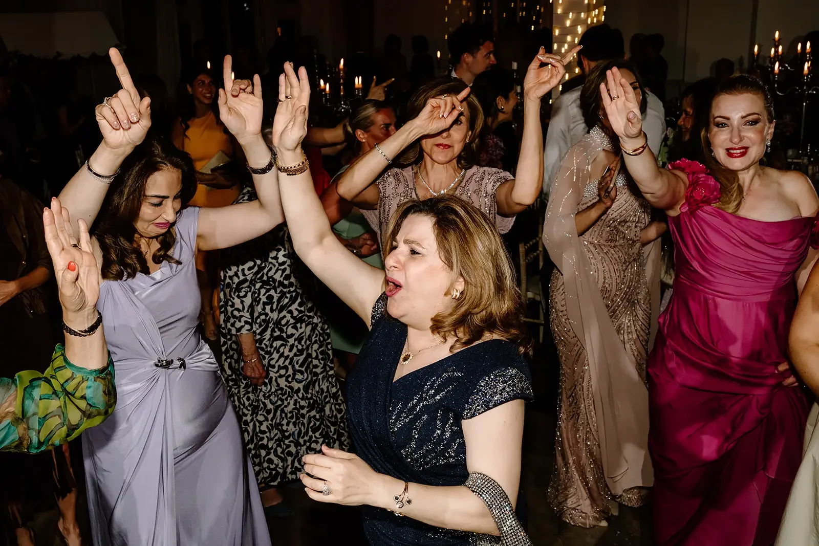 Women dancing and enjoying a celebration at a formal party or event, dressed in elegant gowns, with some women with their arms raised and eyes closed in the moment.