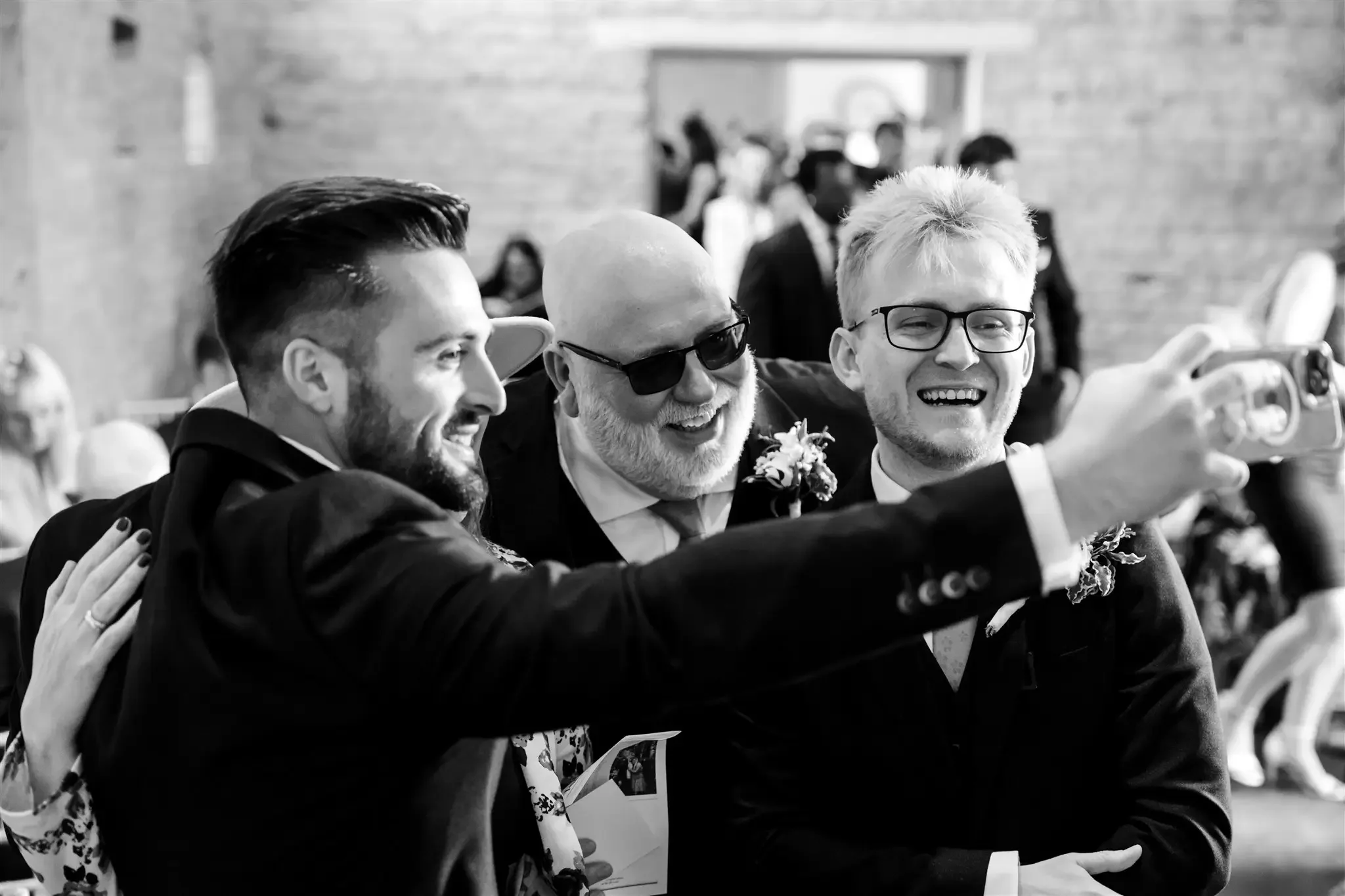 Three men taking a selfie at a wedding, smiling and dressed in formal suits, with a woman in the background and a brick wall.